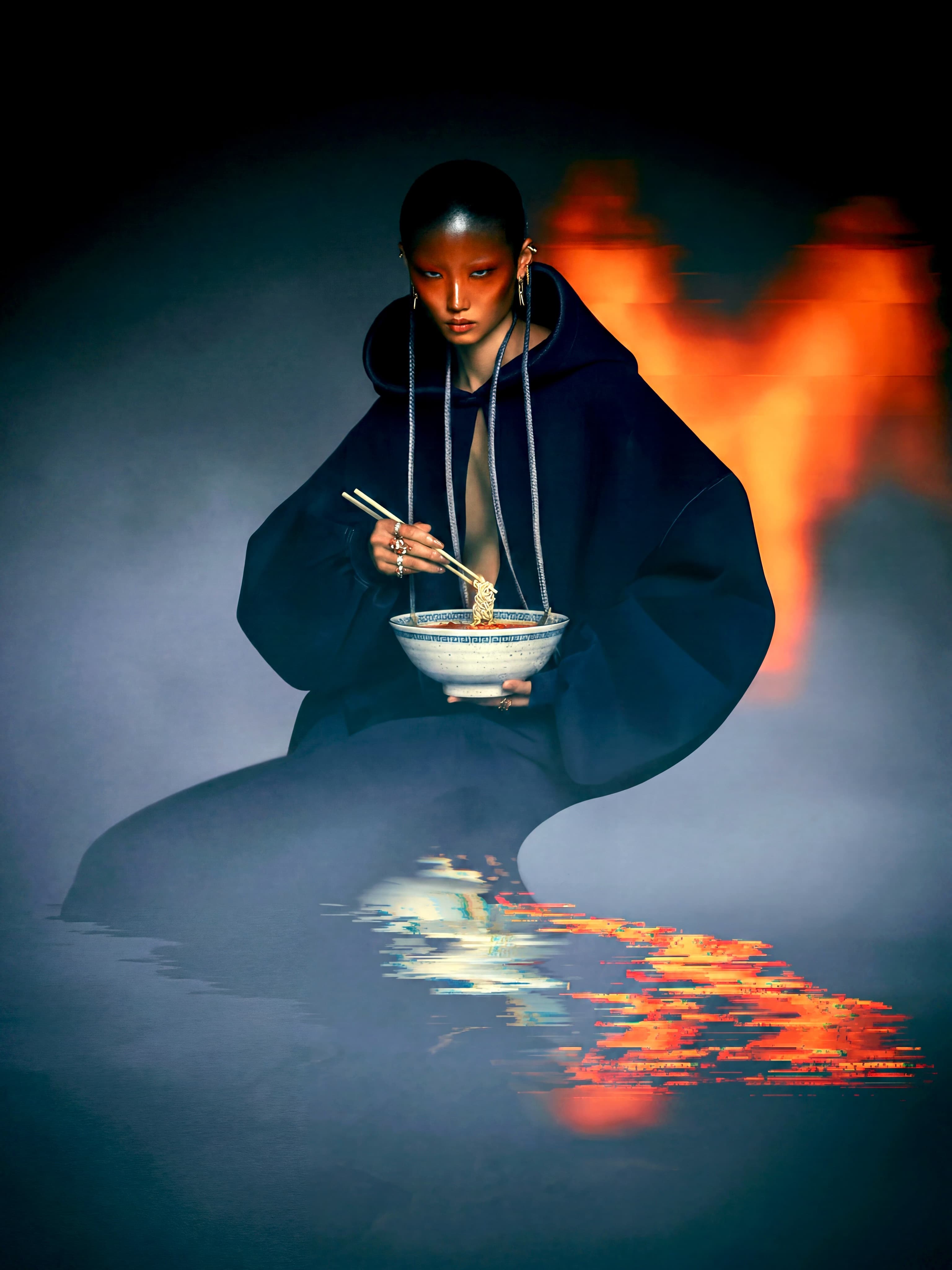 Seated figure in a dark hooded garment holding a bowl and chopsticks before a glowing orange backdrop
